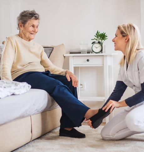 A young health visitor putting on slippers on a senior woman at home at Christmas time.